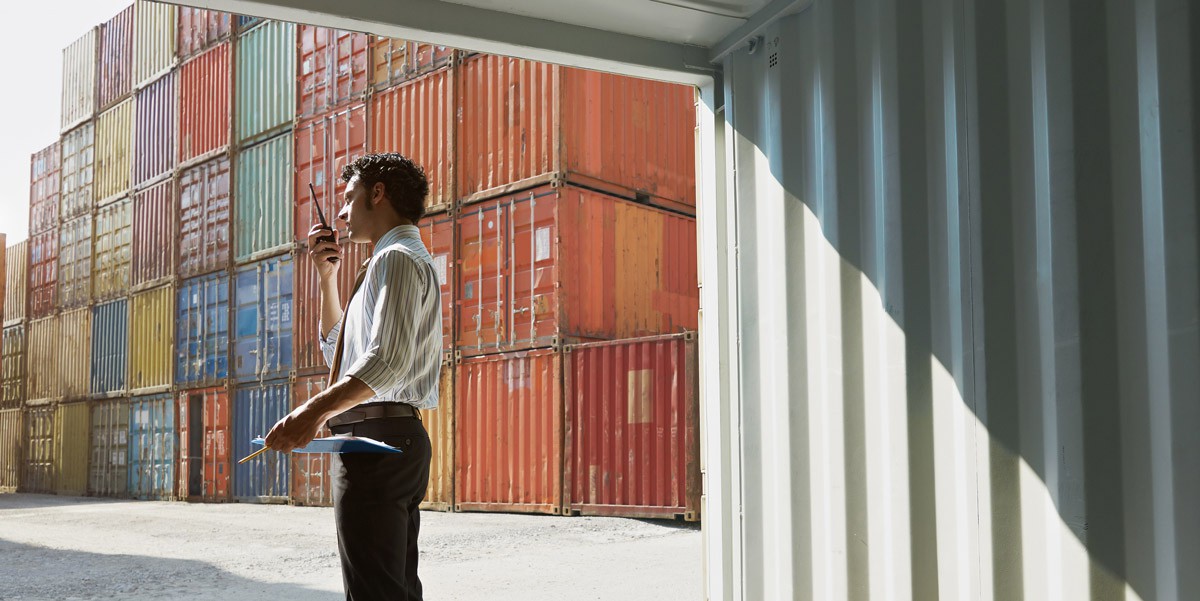 A light-skinned masculine person stands among stacks of cargo containers, talking into a walkie talkie.